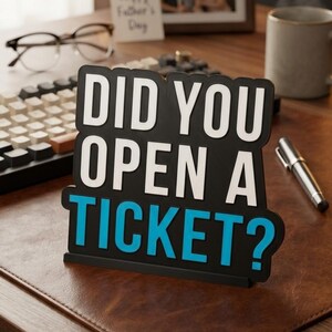Puede incluir: Un letrero negro y azul con el texto "DID YOU OPEN A TICKET?" en letras blancas y azules. El letrero est&aacute; sobre un escritorio de cuero marr&oacute;n con un teclado, gafas, un bol&iacute;grafo y una taza en el fondo. Tambi&eacute;n se ve un marco de fotos y una tarjeta que dice "Happy Father's Day".