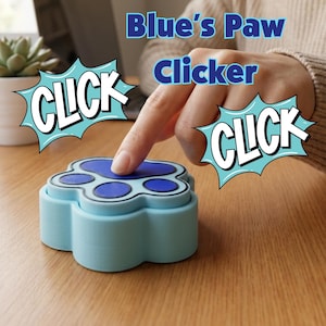 Puede incluir: Un clicker azul claro con forma de pata y un diseño de almohadilla azul oscuro en la parte superior. El dispositivo presenta el texto Blue's Paw Clicker y la palabra CLICK en dos burbujas de estilo cómic. El botón es presionado por un dedo.