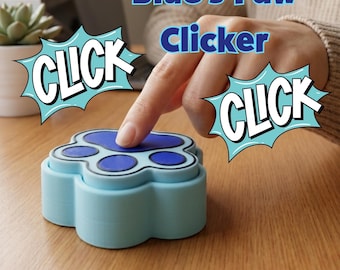 Puede incluir: Un clicker azul claro con forma de pata y un diseño de almohadilla azul oscuro en la parte superior. El dispositivo presenta el texto Blue's Paw Clicker y la palabra CLICK en dos burbujas de estilo cómic. El botón es presionado por un dedo.