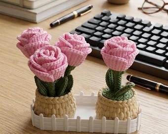 Puede incluir: Arreglo decorativo con cuatro rosas rosas de ganchillo en macetas beige tejidas, colocadas sobre una bandeja blanca con diseño de valla. Las flores tienen tallos y hojas verdes. Sobre un escritorio de madera.
