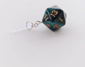Puede incluir: Un dije de dado poliédrico verde azulado y dorado con una cadena plateada y una punta de auricular transparente. El dado tiene números dorados y está conectado a la cadena con un pequeño anillo plateado. El fondo es borroso, verde y blanco.