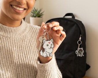 Puede incluir: Un llavero de lobo blanco con un corazón rojo en la frente, sujeto a llaves. El llavero se sostiene frente a una mochila negra. Un segundo llavero con un diseño de gato negro cuelga de la cremallera de la mochila.
