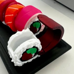 Pode incluir: Um conjunto de rolinhos de sushi coloridos feitos de plástico. Os rolinhos estão dispostos em uma bandeja preta. Os rolinhos são decorados com detalhes verdes e vermelhos.
