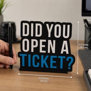 Puede incluir: Un letrero de escritorio negro con el texto "DID YOU OPEN A TICKET?" en blanco y azul. El letrero mide 14 cm de ancho y 10,9 cm de alto. Hay un bol&iacute;grafo negro al lado del letrero.