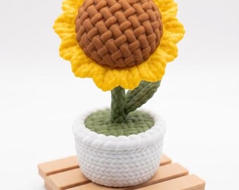Puede incluir: Un girasol de ganchillo hecho a mano en una maceta blanca, sobre una pequeña paleta de madera. El girasol tiene pétalos amarillos y un centro marrón. El tallo es verde.