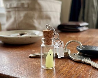 Puede incluir: Una pequeña botella de vidrio con un tapón de corcho, que contiene un dije en forma de fantasma amarillo. La botella está unida a un llavero con un dije rectangular transparente. Llaves sobre una superficie de madera.