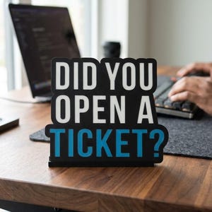 May include: A black and white sign on a wooden desk reads "DID YOU OPEN A TICKET?" in white and blue text. A laptop and keyboard are visible in the background, suggesting a workspace environment.