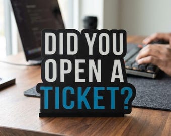 Puede incluir: Un letrero en blanco y negro sobre un escritorio de madera dice "DID YOU OPEN A TICKET?" en texto blanco y azul. Una computadora portátil y un teclado son visibles en el fondo, lo que sugiere un entorno de trabajo.