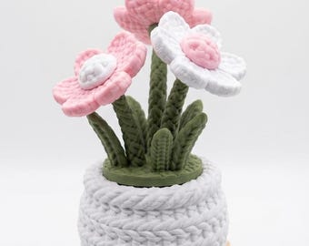 Puede incluir: Arreglo floral de ganchillo en una maceta blanca. Las flores son rosas y blancas con tallos y hojas verdes. La maceta se asienta sobre una pequeña paleta de madera, creando una pieza decorativa encantadora.