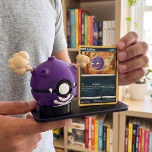 Puede incluir: Una figura de Koffing morada con una tarjeta a juego. La figura tiene un cuerpo redondo con un diseño de calavera blanca y dos nubes de humo beige. La tarjeta es amarilla con el nombre "Koffing" y una imagen del personaje.