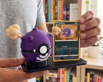Puede incluir: Una figura de Koffing morada con una tarjeta a juego. La figura tiene un cuerpo redondo con un diseño de calavera blanca y dos nubes de humo beige. La tarjeta es amarilla con el nombre "Koffing" y una imagen del personaje.