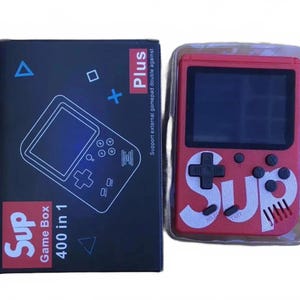 Puede incluir: Una consola de juegos portátil roja con la palabra "Sup" en blanco. La consola tiene una pantalla, una cruceta y botones. La caja de al lado es negra con un diagrama de la consola y las palabras "Sup Game Box 400 in 1".