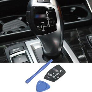 May include: A silver and black car gear shift lever with the letters R, N, D, and P. A blue plastic pry tool and a black gear shift panel are also visible.