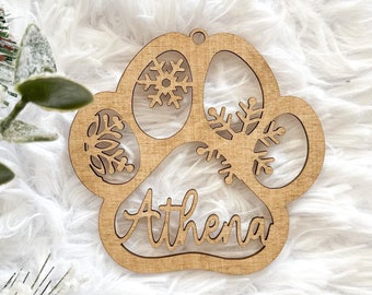 May include: A light brown wooden ornament shaped like a paw print featuring cut-out snowflake designs and the name Athena written in cursive script across the center. The ornament includes a small hole at the top for hanging purposes.