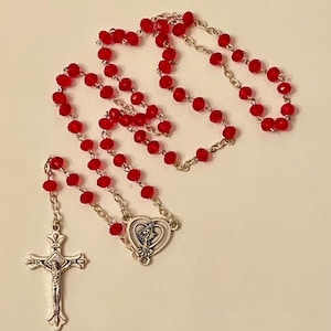 May include: A red crystal rosary with a silver heart charm and a silver crucifix. The crucifix features a depiction of Jesus Christ.