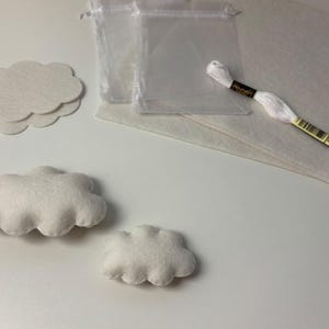 Könnte beinhalten: Ein Bastelset mit weißen Wolkenformen aus Filz, zwei transparenten Beuteln mit Kordelzug, einem rechteckigen Stück weißem Filz und einem Strang weißem Stickgarn. Das Set enthält sowohl flache als auch gefüllte, dreidimensionale Wolken.