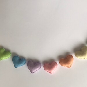 May include: A garland of felt hearts in a rainbow of pastel colors, including pink, orange, yellow, green, blue, and purple. Each heart is hand-stitched with a contrasting thread, creating a charming and decorative accent.