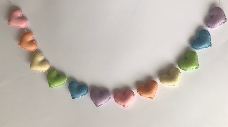 May include: A garland of felt hearts in a rainbow of pastel colors, including pink, orange, yellow, green, blue, and purple. Each heart is hand-stitched with a contrasting thread, creating a charming and decorative accent.