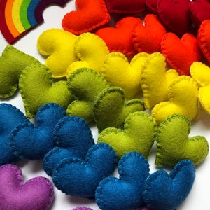 May include: A collection of colorful felt hearts in a rainbow of colors, including red, orange, yellow, green, blue, and purple. A felt rainbow is also present. The hearts are hand-stitched with contrasting thread, creating a charming, handmade aesthetic.