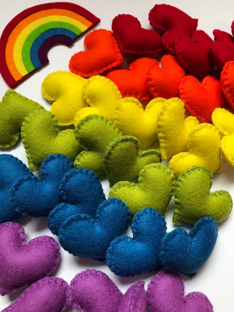 May include: A collection of colorful felt hearts in a rainbow of colors, including red, orange, yellow, green, blue, and purple. A felt rainbow is also present. The hearts are hand-stitched with contrasting thread, creating a charming, handmade aesthetic.