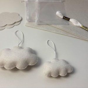 Op de afbeelding: Een knutselset met twee afgewerkte witte vilten wolkornamenten met ophanglussen, drie platte witte vilten wolkuitsparingen, twee doorzichtige plastic zakjes, een vel wit vilt en een streng wit borduurgaren voor de montage.