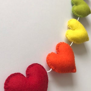 May include: A colorful felt heart garland in a rainbow of colors, including red, orange, yellow, green, blue, and purple. Each heart is hand-stitched with white thread and strung on a white cord. Perfect for festive decor.