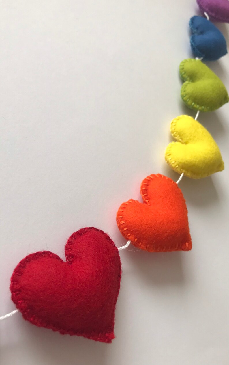 May include: A colorful felt heart garland in a rainbow of colors, including red, orange, yellow, green, blue, and purple. Each heart is hand-stitched with white thread and strung on a white cord. Perfect for festive decor.