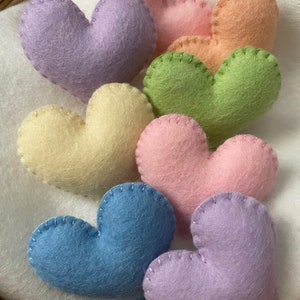 Könnte beinhalten: Sieben Filz-Herzen in Pastellfarben: Lavendel, Hellgelb, Hellgrün, Rosa, Hellblau und Lila. Die Herzen liegen auf einer weißen Oberfläche.