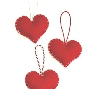 May include: Three red felt heart ornaments with white stitching and hanging loops. The hearts have a scalloped edge and are suspended by white or red and white twine. Perfect for Valentine's Day or other celebrations.