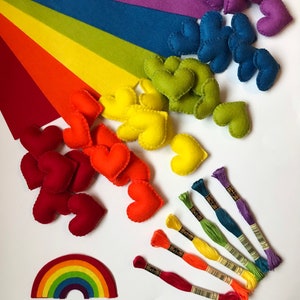 May include: A collection of colorful craft supplies arranged on a white surface. The image features felt hearts in various colors, rainbow-colored embroidery floss, and felt rainbow shapes. The color palette includes red, orange, yellow, green, blue, and purple.