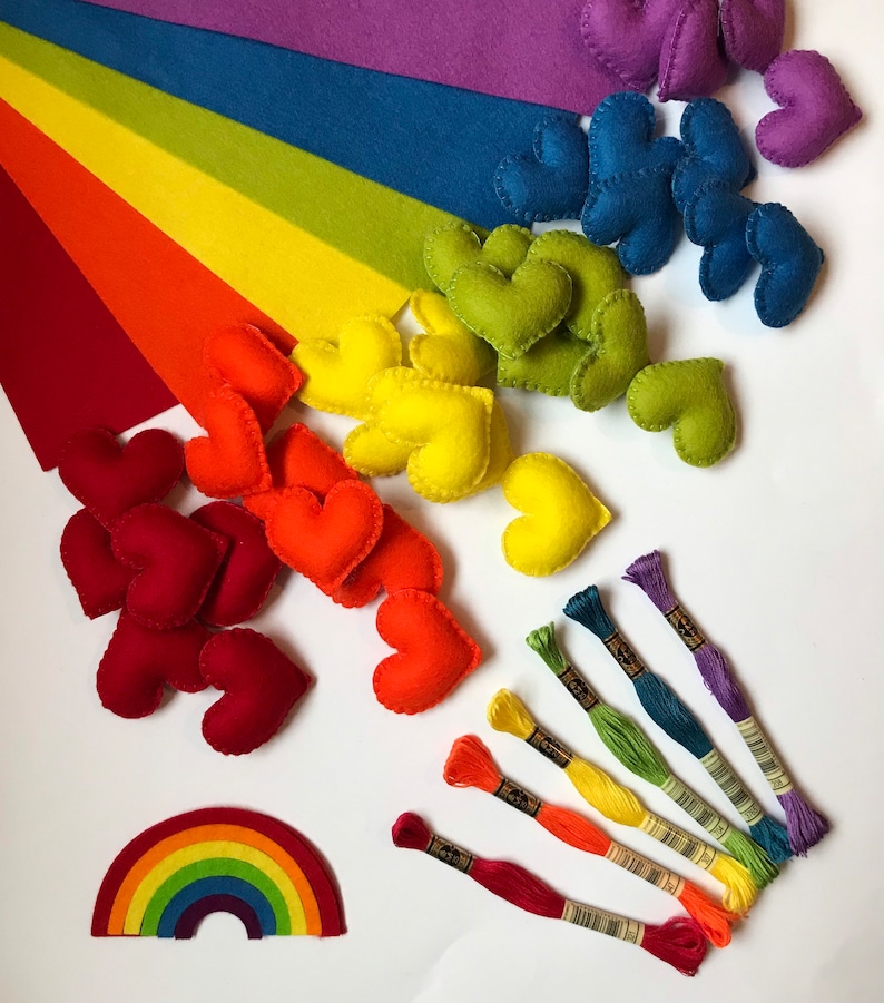 May include: A collection of colorful craft supplies arranged on a white surface. The image features felt hearts in various colors, rainbow-colored embroidery floss, and felt rainbow shapes. The color palette includes red, orange, yellow, green, blue, and purple.