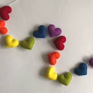 May include: A colorful heart garland featuring felt hearts in a rainbow of colors: red, orange, yellow, green, blue, and purple. The hearts are strung together with white thread, creating a cheerful and decorative accent.
