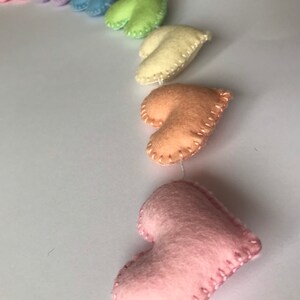 May include: A garland of felt hearts in a rainbow of pastel colors: pink, lavender, blue, green, yellow, and peach. Each heart is hand-stitched with contrasting thread and strung together with white string, creating a charming decorative item.