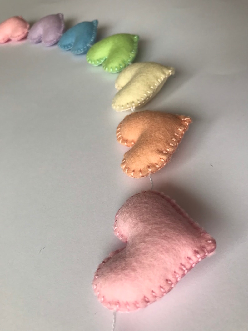 May include: A garland of felt hearts in a rainbow of pastel colors: pink, lavender, blue, green, yellow, and peach. Each heart is hand-stitched with contrasting thread and strung together with white string, creating a charming decorative item.