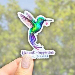 May include: A sticker featuring a vibrant hummingbird with green, blue, and purple hues, holding a pink flower in its beak. The sticker has the text "Eternal Happiness" and "Ecuador" in a purple font.