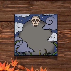 May include: A spooky Halloween card with a cartoon skull peeking out from behind a gray rock. The background is a dark blue night sky with white clouds and stars.