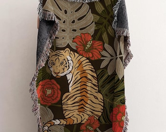 May include: A woven throw blanket featuring a tiger illustration surrounded by tropical flowers and leaves in shades of orange, red, green, and brown. The blanket has a fringed edge and a dark gray backing.
