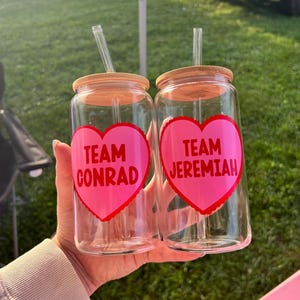 May include: Two clear glass tumblers with bamboo lids and clear straws. Each tumbler has a pink heart with red trim and text. One says "Team Conrad" and the other says "Team Jeremiah".