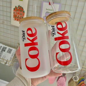 May include: Two glass tumblers with bamboo lids and red straws. Both tumblers have a white frosted finish and feature the words "Diet Coke" in red lettering.