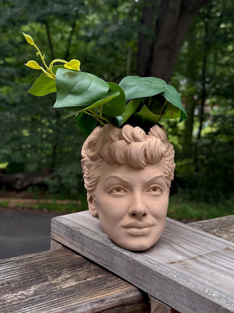 May include: A beige-colored planter shaped like a woman's head, with green leafy plants sprouting from the top. The planter is made of a solid material and sits on a wooden surface, with a blurred green background.