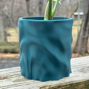 Op de afbeelding: Teal gekleurde, getextureerde plantenpot met een golvend ontwerp. De cilindrische pot bevat een groene plant met lange, slanke bladeren. De pot staat op een verweerd houten oppervlak, met een vage achtergrond.