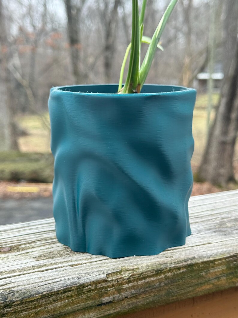 Op de afbeelding: Teal gekleurde, getextureerde plantenpot met een golvend ontwerp. De cilindrische pot bevat een groene plant met lange, slanke bladeren. De pot staat op een verweerd houten oppervlak, met een vage achtergrond.