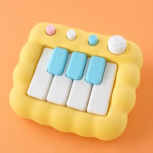 May include: A yellow toy piano with white and blue keys. The toy has a scalloped edge and several colored buttons and a white knob. The background is a solid orange color.