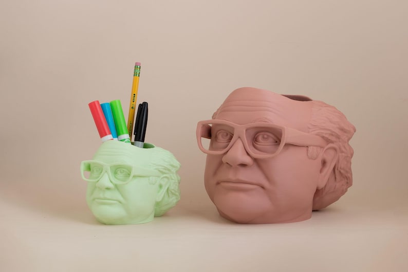 Op de afbeelding: Twee bureau-organizers in de vorm van een mannenhoofd met een bril. E&eacute;n is groen en de andere is roze. De groene organizer heeft een potlood en stiften erin.