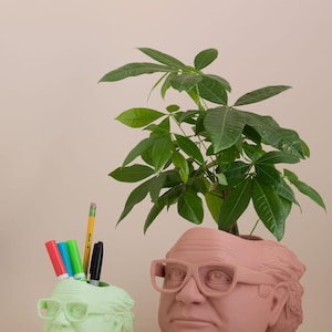 Op de afbeelding: Twee keramische potten in de vorm van een mensenhoofd, een groen en een bruin. De groene pot heeft een potlood en stiften erin. De bruine pot heeft een kleine groene plant die uit de bovenkant groeit.