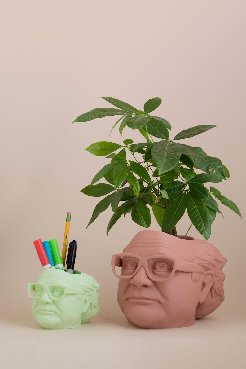 Op de afbeelding: Twee keramische potten in de vorm van een mensenhoofd, een groen en een bruin. De groene pot heeft een potlood en stiften erin. De bruine pot heeft een kleine groene plant die uit de bovenkant groeit.