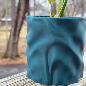 Op de afbeelding: Teal gekleurde, getextureerde plantenpot met een uniek, gekreukeld ontwerp. De pot bevat een kleine plant met groene bladeren. De pot staat op een houten oppervlak, met een vage achtergrond.