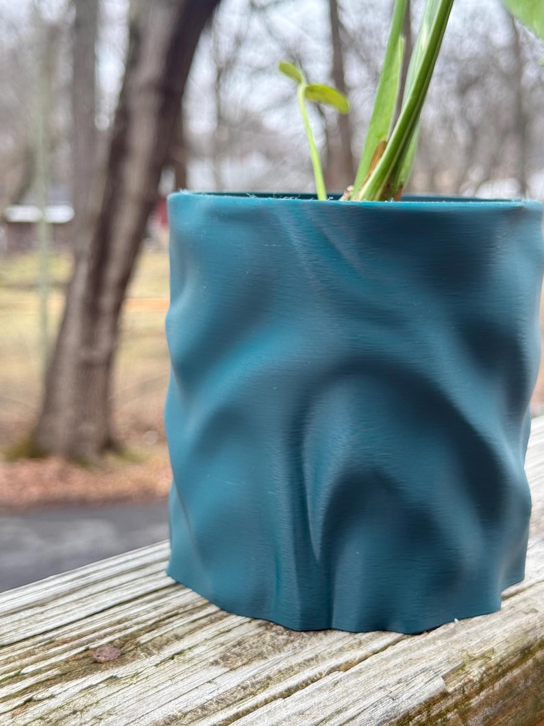 Op de afbeelding: Teal gekleurde, getextureerde plantenpot met een uniek, gekreukeld ontwerp. De pot bevat een kleine plant met groene bladeren. De pot staat op een houten oppervlak, met een vage achtergrond.