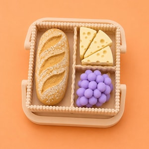 May include: A beige divided tray filled with a loaf of bread, three cheese wedges, and a cluster of purple grapes. The tray has a beaded border and is set against an orange background. The bread is golden brown with a textured surface.