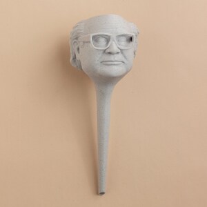 May include: A white ceramic sculpture of a person's head with glasses. The sculpture is attached to a long, thin, white stem.